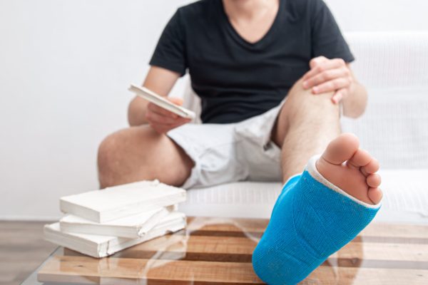 Close up man’s broken leg in blue splint for treatment of injuries from ankle sprain at home. Man with broken leg in blue splint for treatment of injuries from ankle sprain reading books at the home rehabilitation.